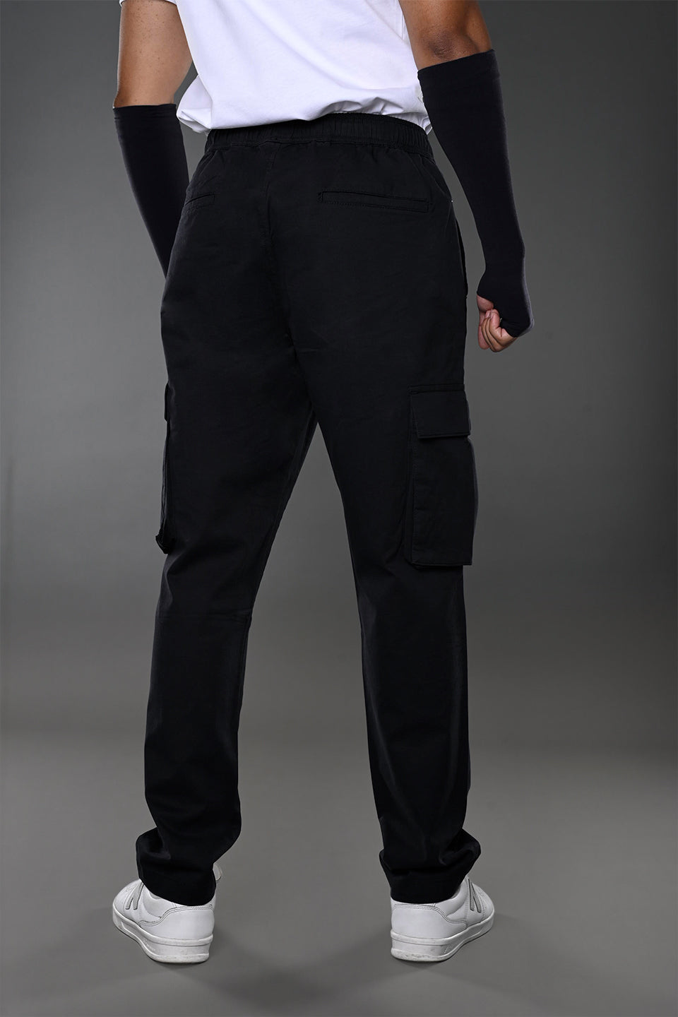 Back view of man in white t-shirt, black UNDERRATED CLUB cargo pants with side pockets, wristbands, and white sneakers.