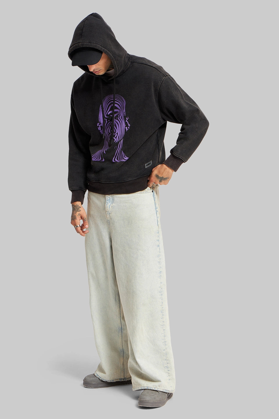 Full-body view of a person wearing a black hoodie with purple graphics and wide-leg light grey trousers.