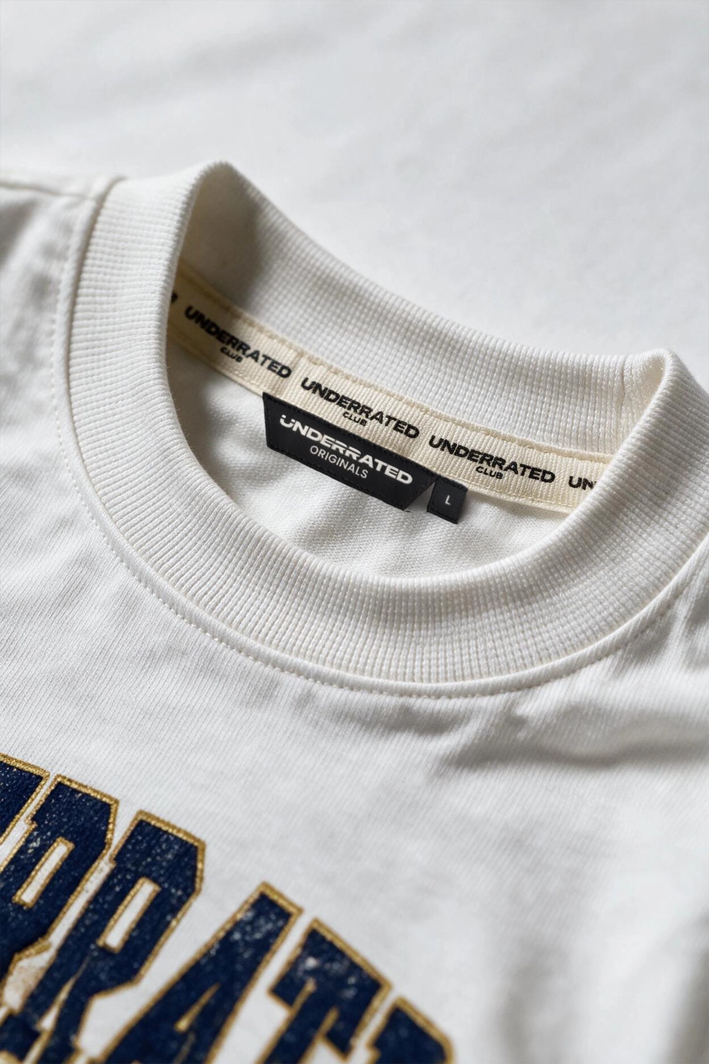 Close-up of a white crewneck t-shirt showing the inner neck label with 'UNDERRAATED' branding and the top of a navy blue and gold graphic print.