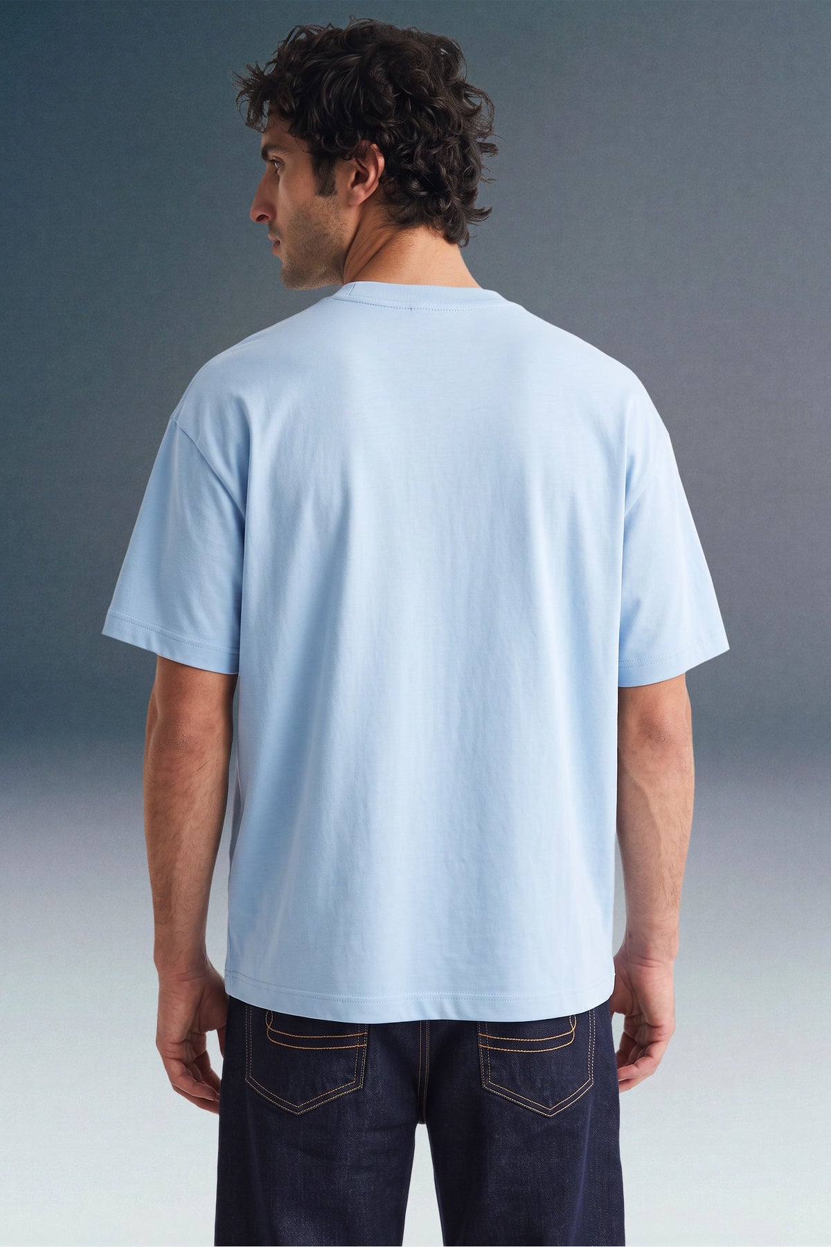 Rear view from the waist up of a man looking to his right, showcasing the back fit and relaxed sleeves of a light blue crewneck T-shirt paired with dark blue jeans.