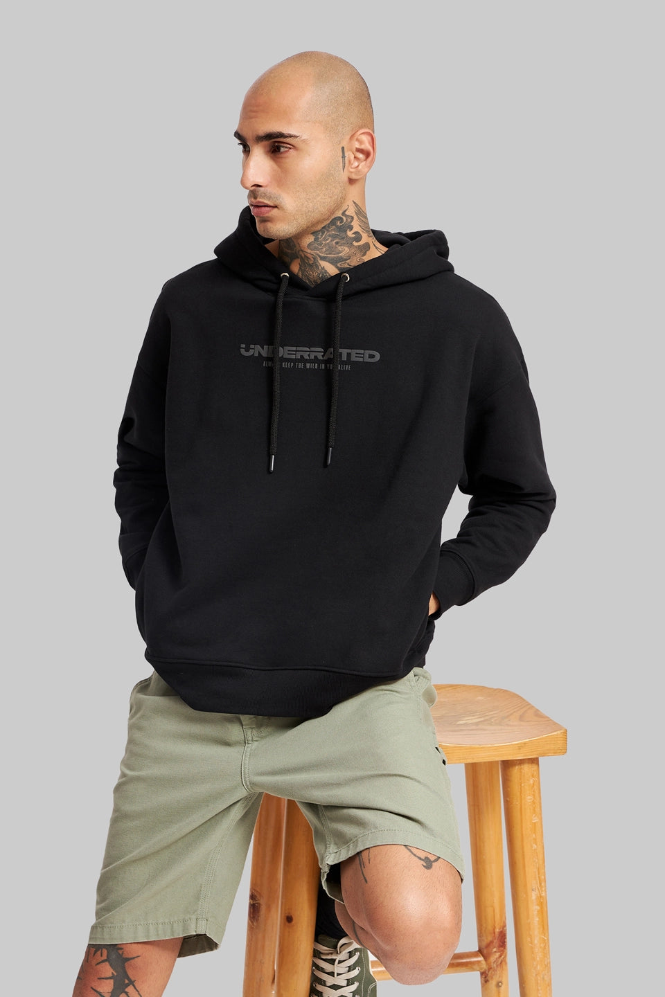 A man in a black "Underrated Club" hoodie and olive green shorts sitting on a wooden stool.