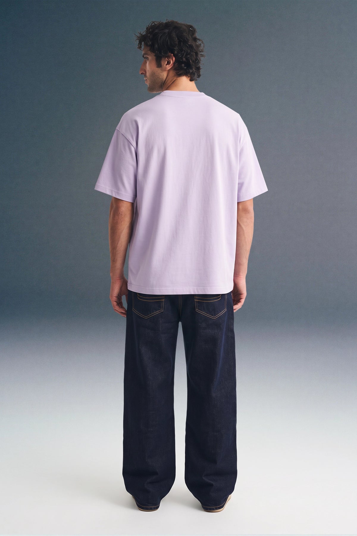 Full-length rear view of a man standing straight, showing the back profile of a light purple T-shirt paired with dark blue trousers.