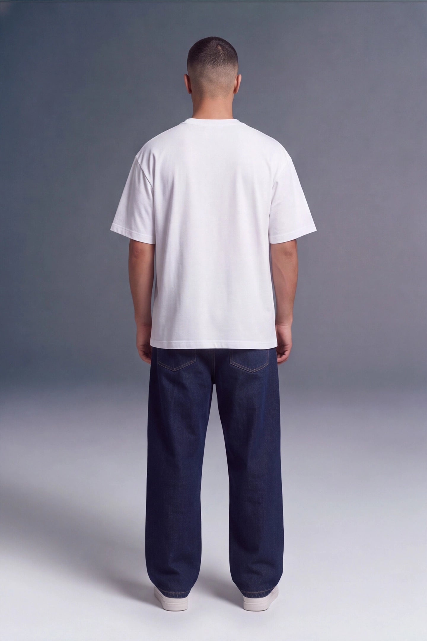 A man viewed from behind wearing a plain white short-sleeve t-shirt and dark navy blue jeans, standing against a gray studio background.