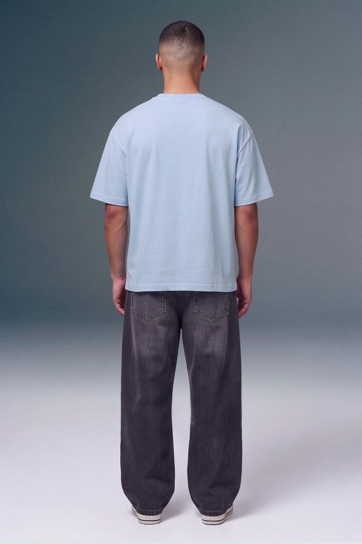 A man viewed from behind wearing a light blue short-sleeve t-shirt and dark gray relaxed-fit jeans, standing against a studio background with a gray-to-white gradient.