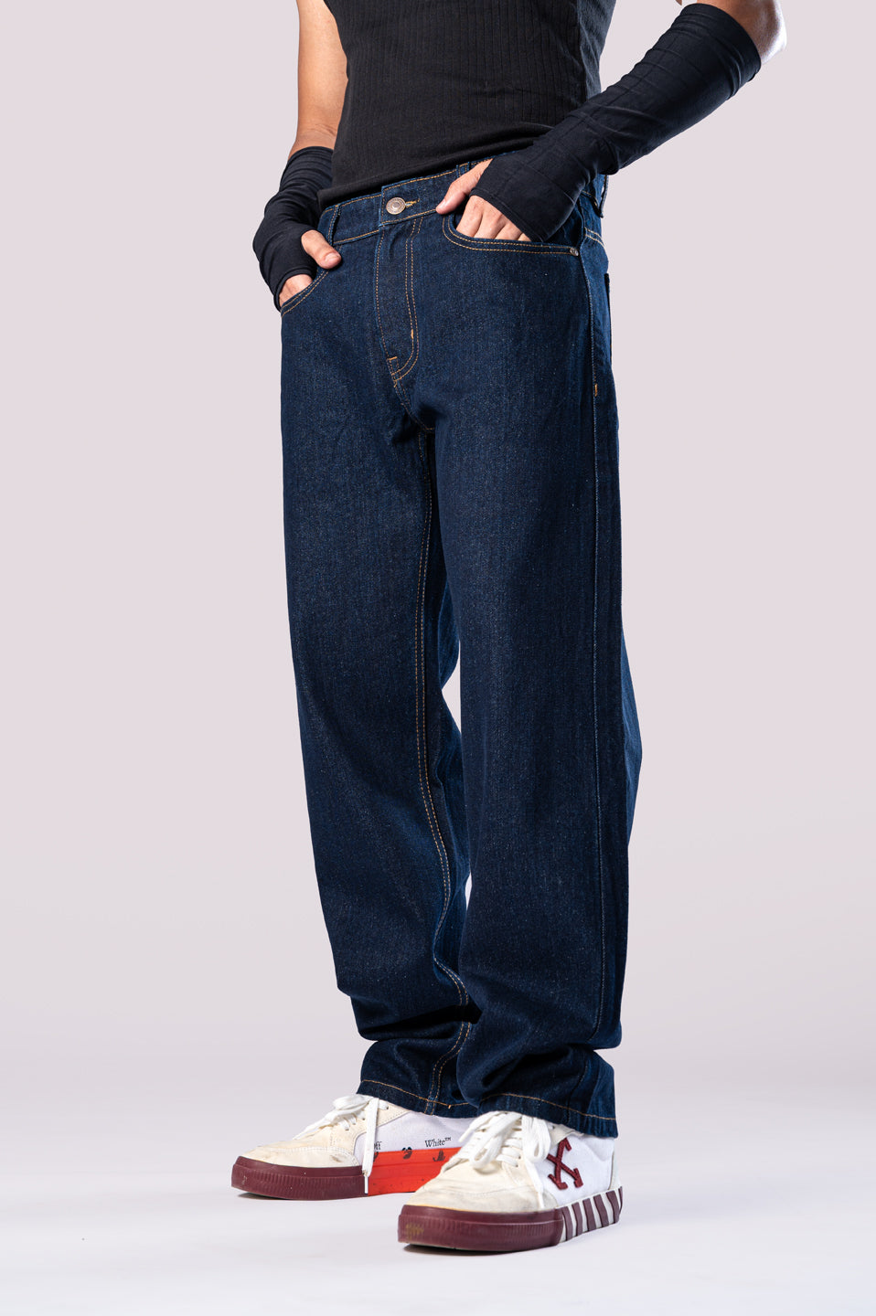 Side view of a person wearing dark-wash blue baggy jeans, a black ribbed top, black arm sleeves, and white and maroon sneakers, with their hands in their pockets.