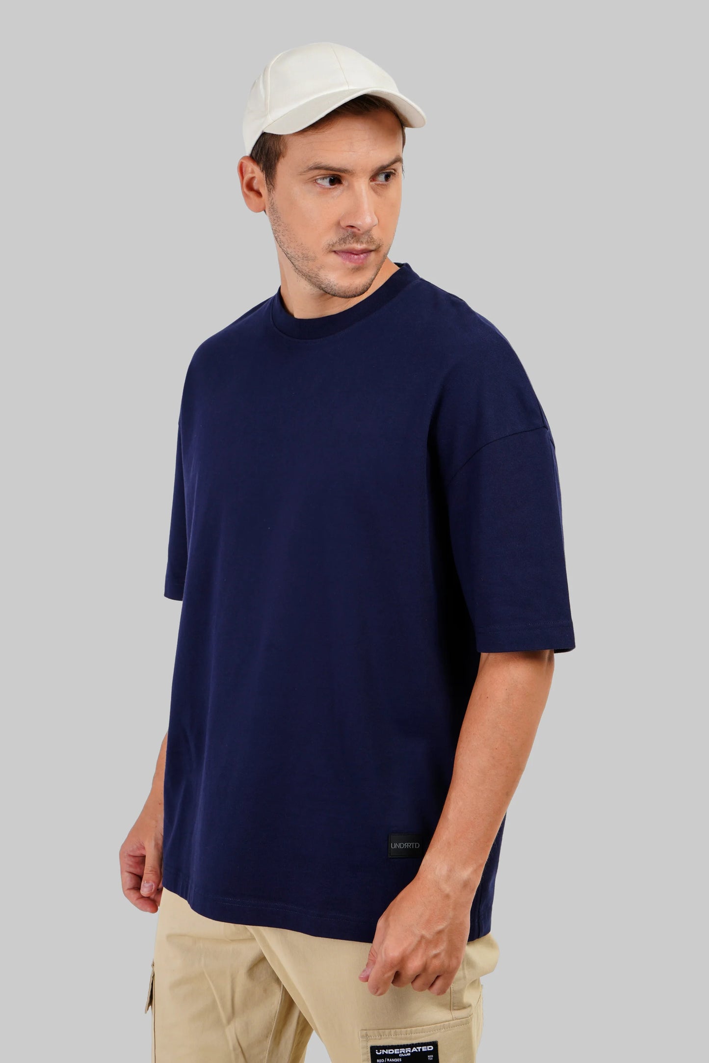 A man wearing an oversized navy blue crewneck t-shirt, beige cargo pants, and a white baseball cap, photographed against a solid gray studio background.