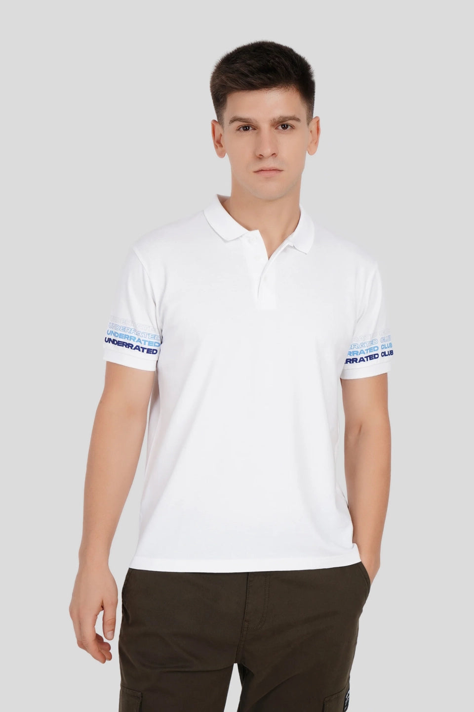 A man wearing a white polo shirt with blue striped cuffs and dark trousers against a neutral grey background.
