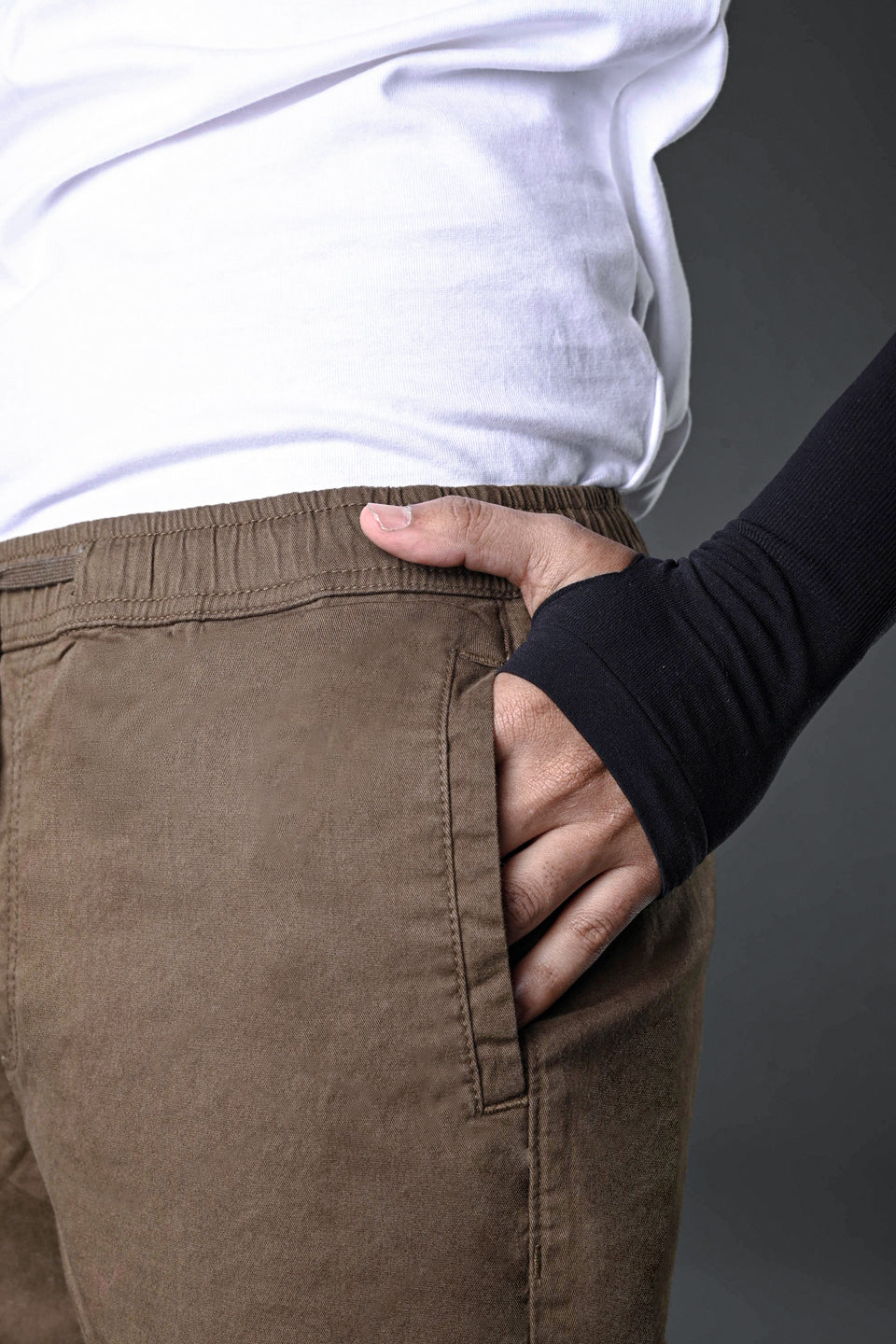 Model hand in waistband of brown UNDERRATED CLUB drawstring cargo pants, white t-shirt, black wristbands against grey background.