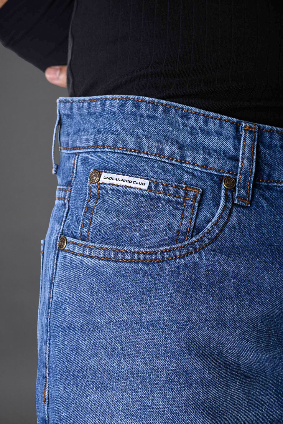 Side angle blue jeans waistband "Underrated" branded leather patch pocket topstitching black top white sneakers.