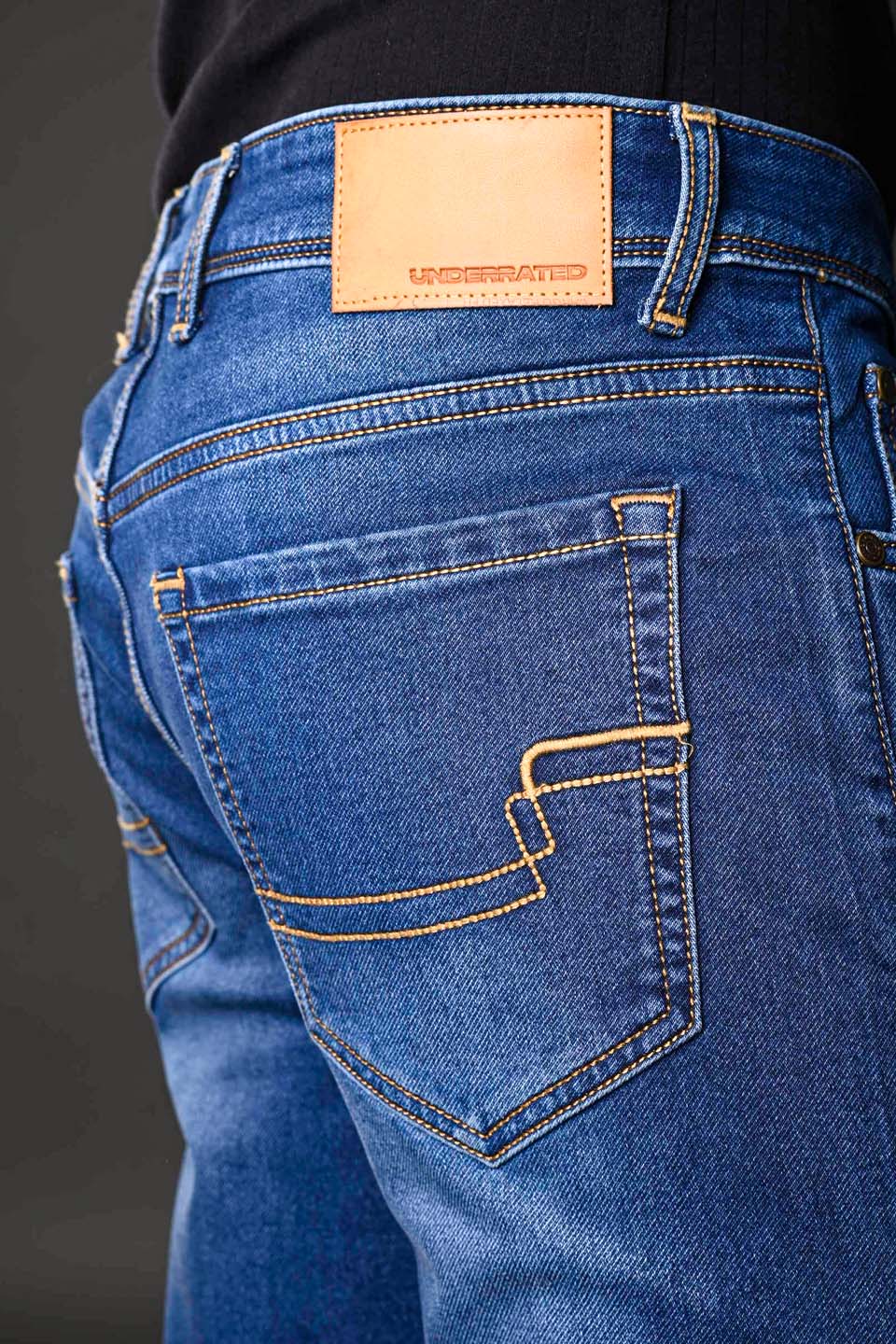 Close-up back pocket Underrated Club leather label tan pocket flap yellow curved stitching detail blue denim jeans.