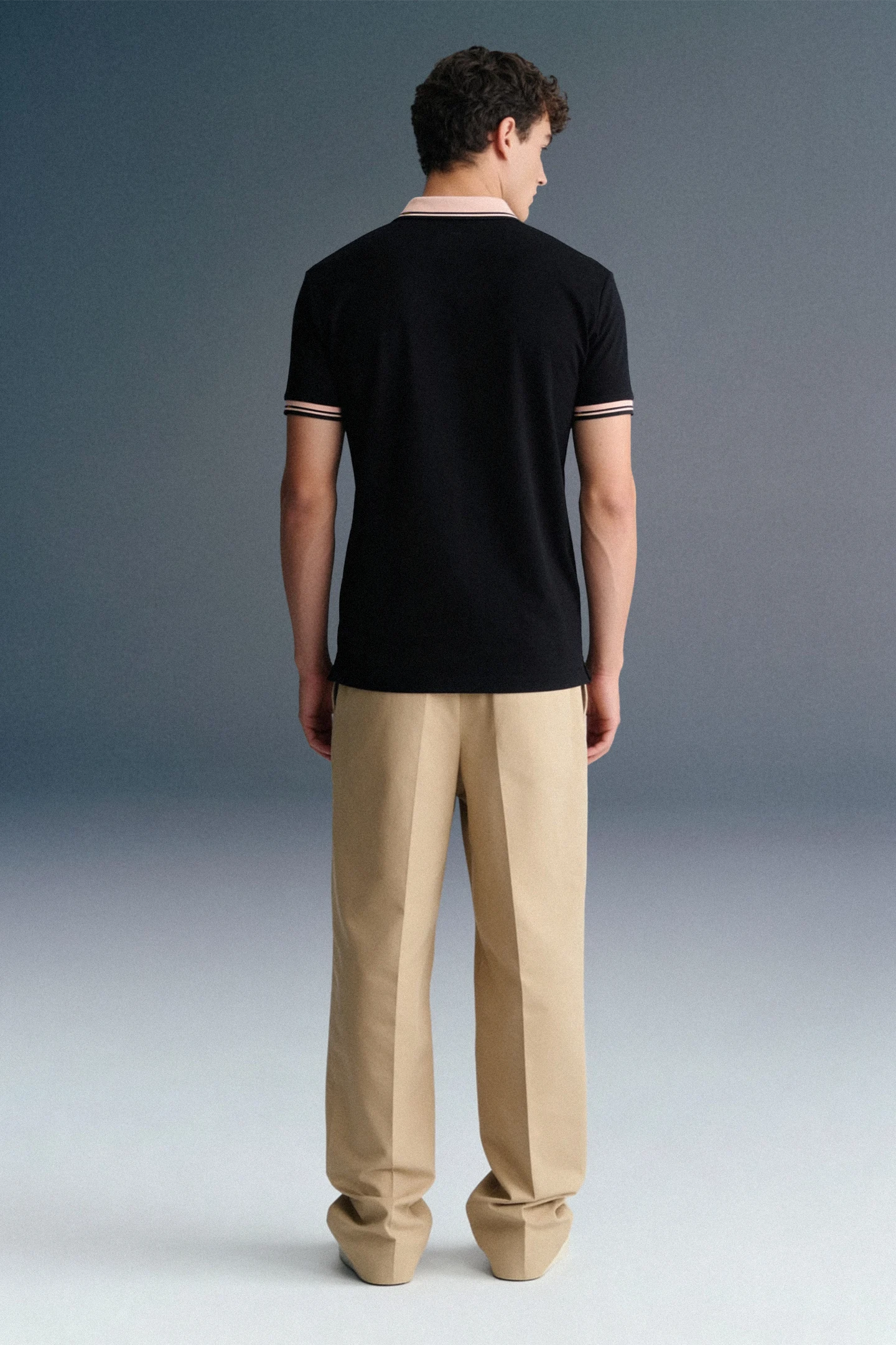 Full-length rear view of a man standing straight, wearing a black polo shirt with a beige collar and patterned sleeve trim, paired with khaki trousers.