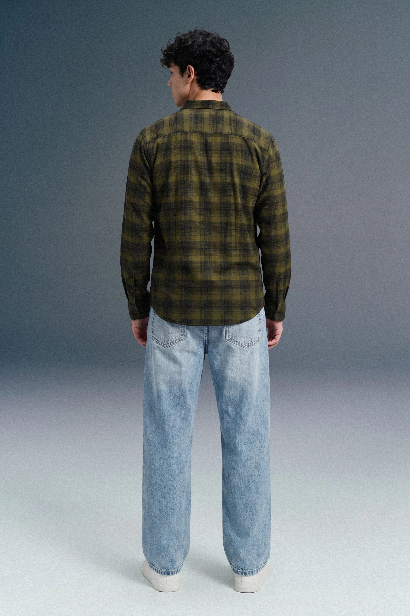 Person wearing a green plaid shirt and light blue jeans against a gray background