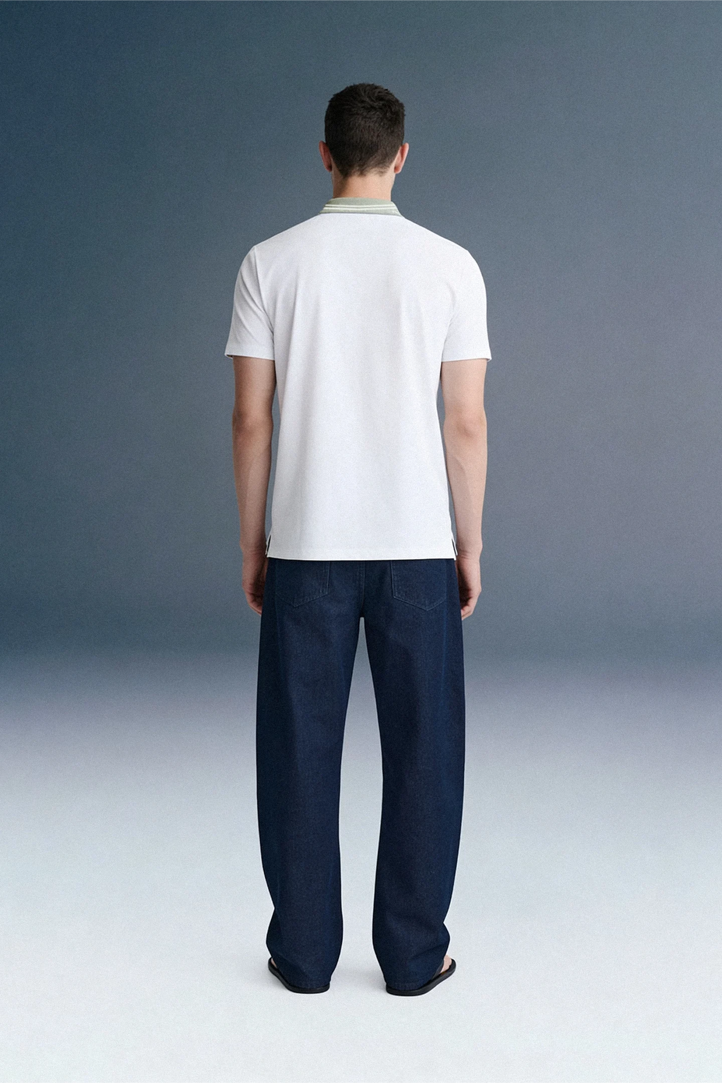 Full-length rear view of a man standing straight, wearing a white polo shirt with an olive green collar and dark navy blue trousers.