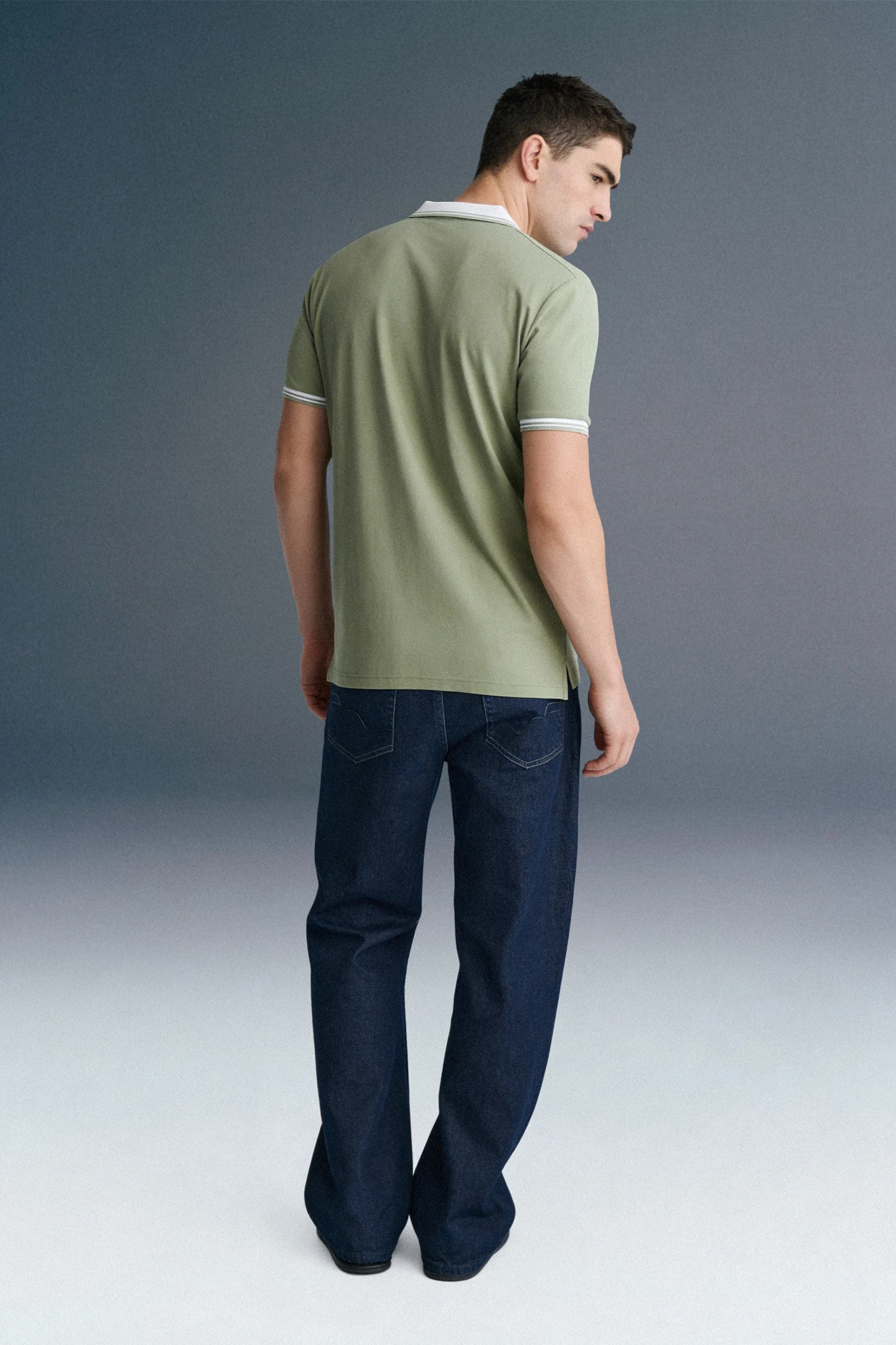 Full-length rear view of a man looking over his shoulder, wearing an olive green polo shirt with a white collar paired with dark navy blue trousers.