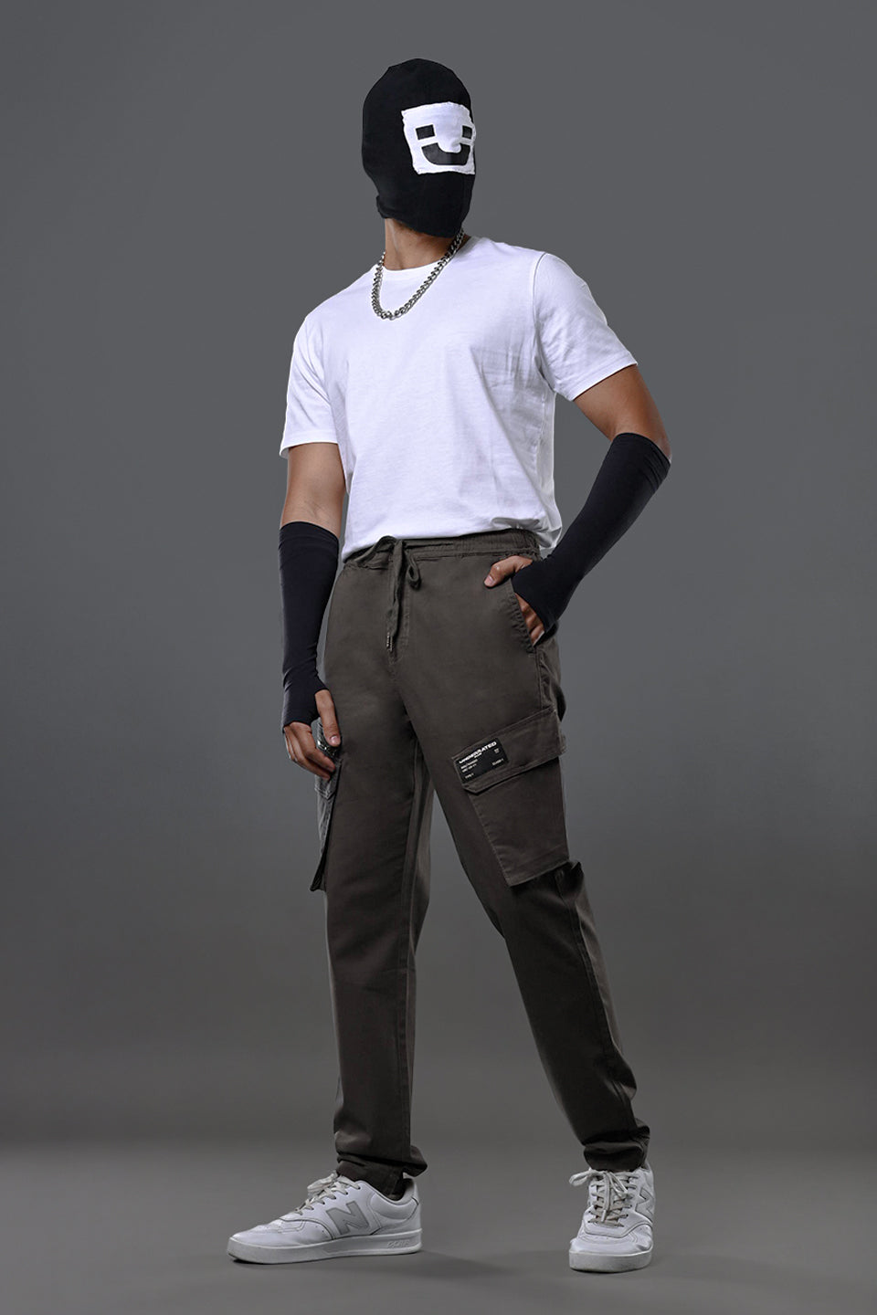 Front view of man wearing black smiley mask, white t-shirt with chain necklace, charcoal grey UNDERRATED cargo pants, wristbands, and white sneakers posing dynamically.