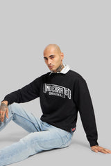 A man sitting in light blue jeans and a black sweatshirt with a bold white graphic across the chest.
