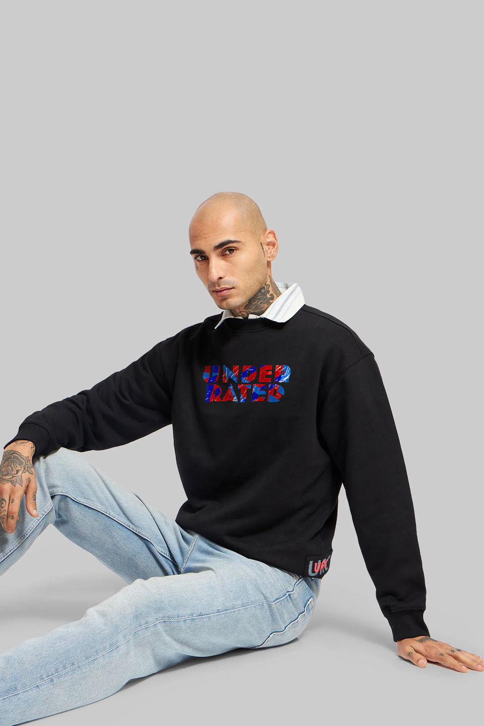 Man seated casually on the floor wearing a black sweatshirt with a colorful red-and-blue “Underrated” graphic, layered over a white collared shirt with light blue jeans on a grey background.