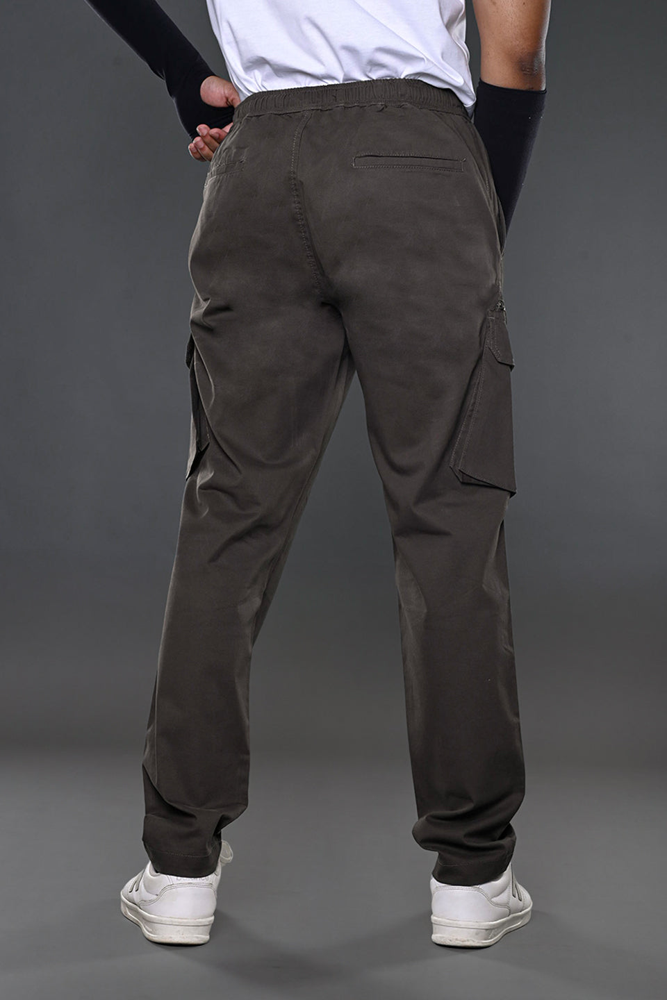 Back view of man in white t-shirt, charcoal grey drawstring UNDERRATED Club cargo pants with side flap pockets, wristbands, and white sneakers against grey background.