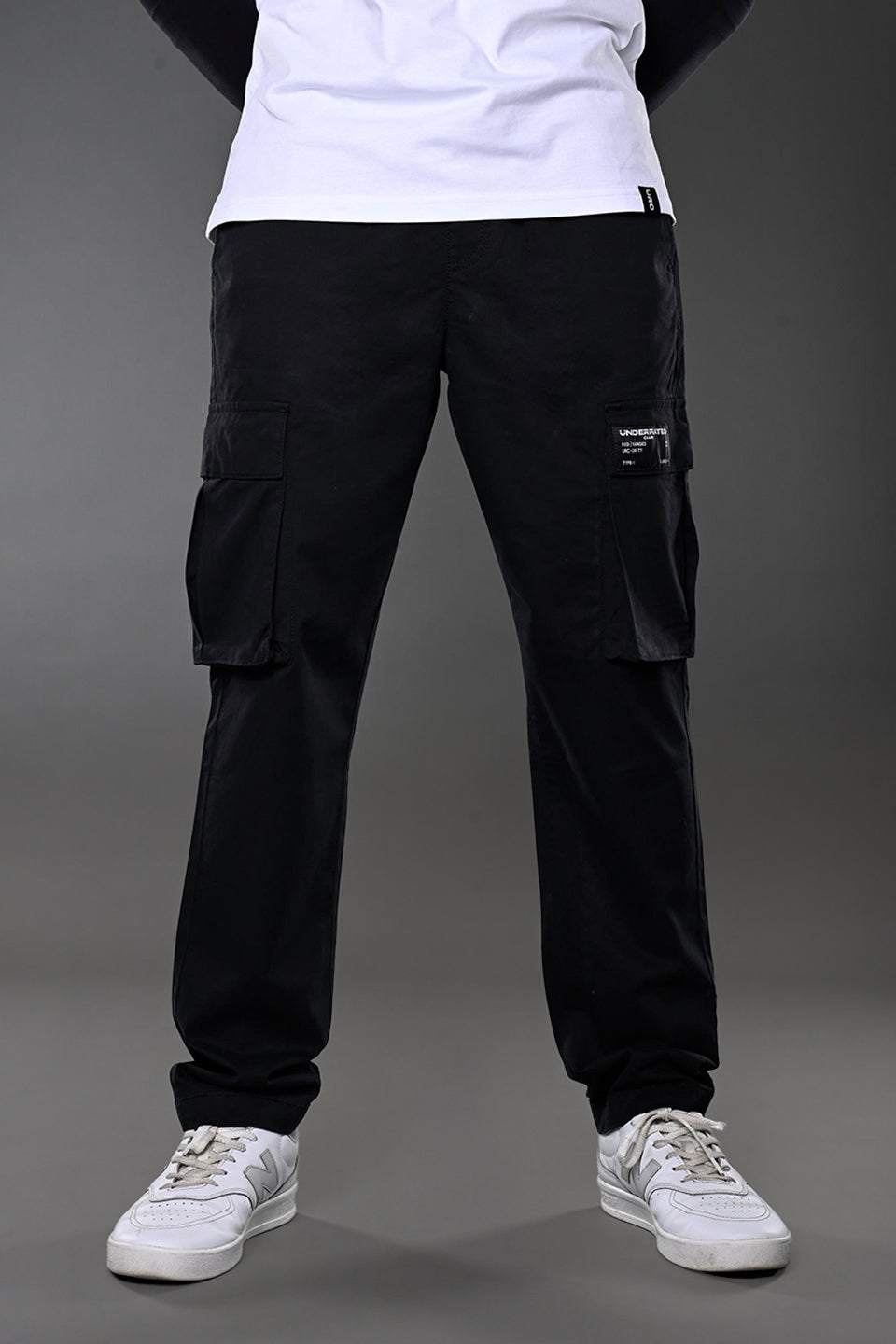 Full-body view of men's black UNDERRATED CLUB cargo pants with flap pockets and label patch, styled with white t-shirt and sneakers.
