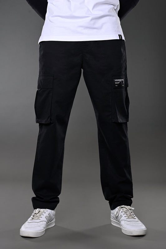 Full-body view of men's black UNDERRATED CLUB cargo pants with flap pockets and label patch, styled with white t-shirt and sneakers.