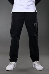 Full-body view of men's black UNDERRATED CLUB cargo pants with flap pockets and label patch, styled with white t-shirt and sneakers.