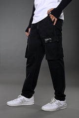 Side view of black UNDERRATED CLUB cargo pants with multiple flap pockets and woven label, styled with white t-shirt, wristbands, and white sneakers.