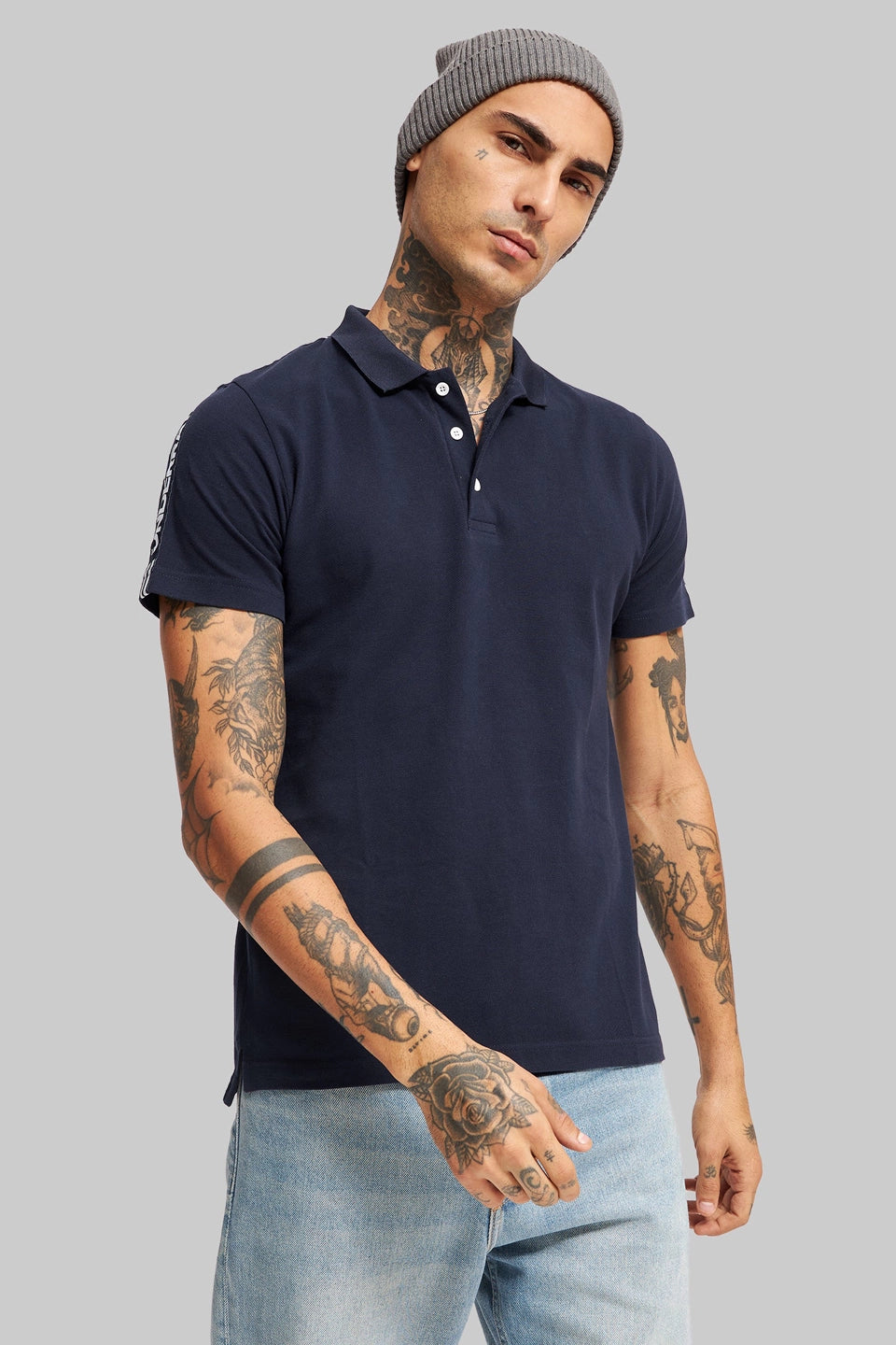 A man with tattoos wearing a navy blue polo shirt and a grey beanie against a plain grey background.