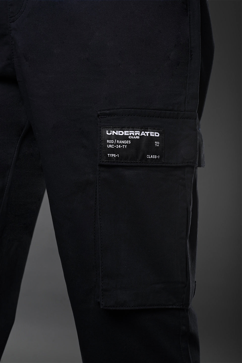 Close-up of black UNDERRATED cargo pants thigh pocket with woven label patch reading "TP-1 D3-1