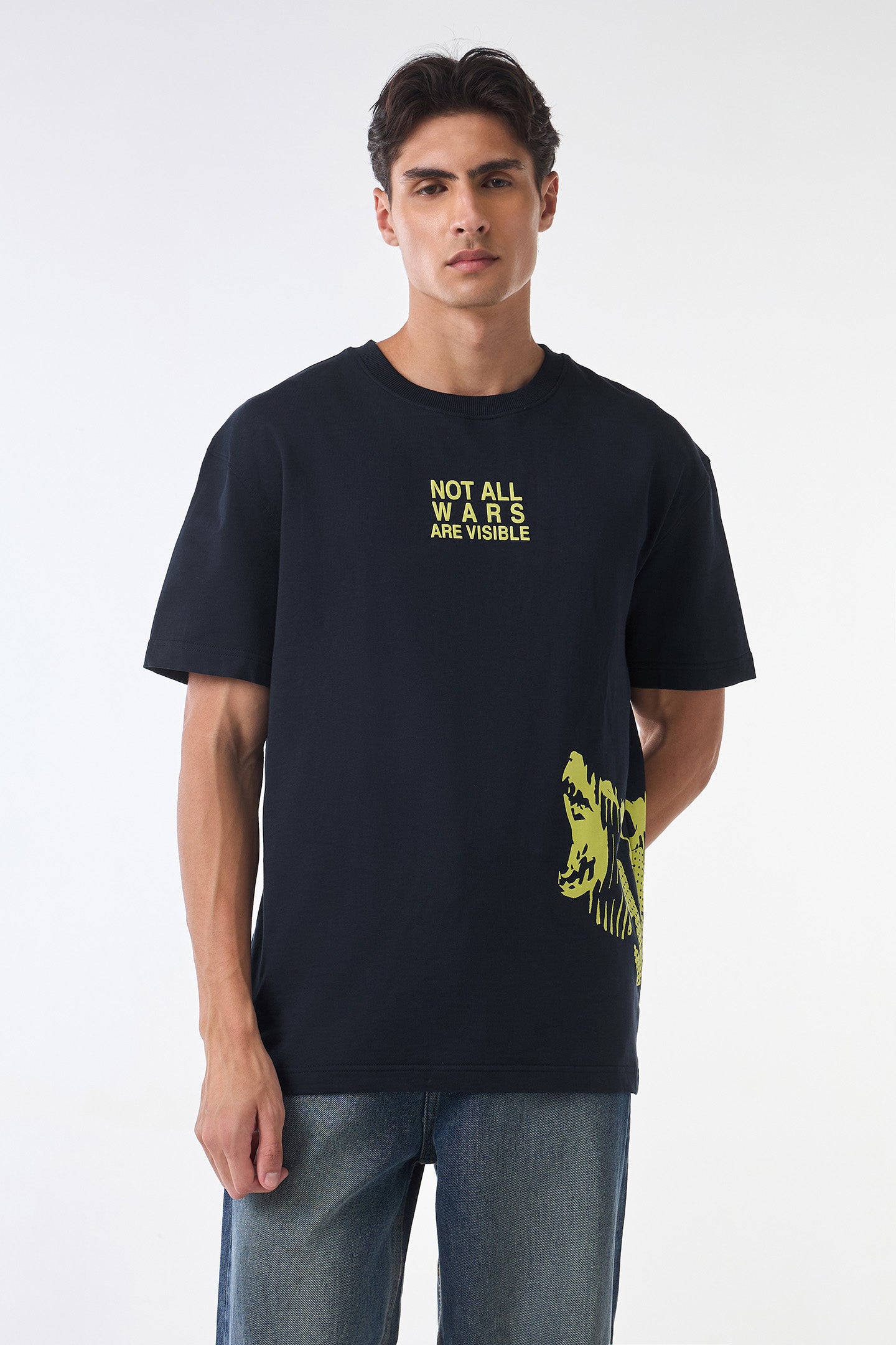 Front studio view of the man standing straight in the “NOT ALL WARS ARE VISIBLE” black t-shirt with yellow side graphic, blue jeans, and neutral background.