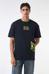 Front studio view of the man standing straight in the “NOT ALL WARS ARE VISIBLE” black t-shirt with yellow side graphic, blue jeans, and neutral background.