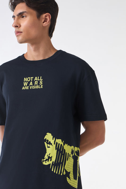 Close-up of a man wearing a black graphic t-shirt with yellow text reading “NOT ALL WARS ARE VISIBLE” on the chest and a bold yellow abstract face print near the hem.
