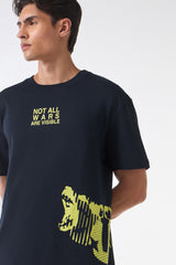 Close-up of a man wearing a black graphic t-shirt with yellow text reading “NOT ALL WARS ARE VISIBLE” on the chest and a bold yellow abstract face print near the hem.