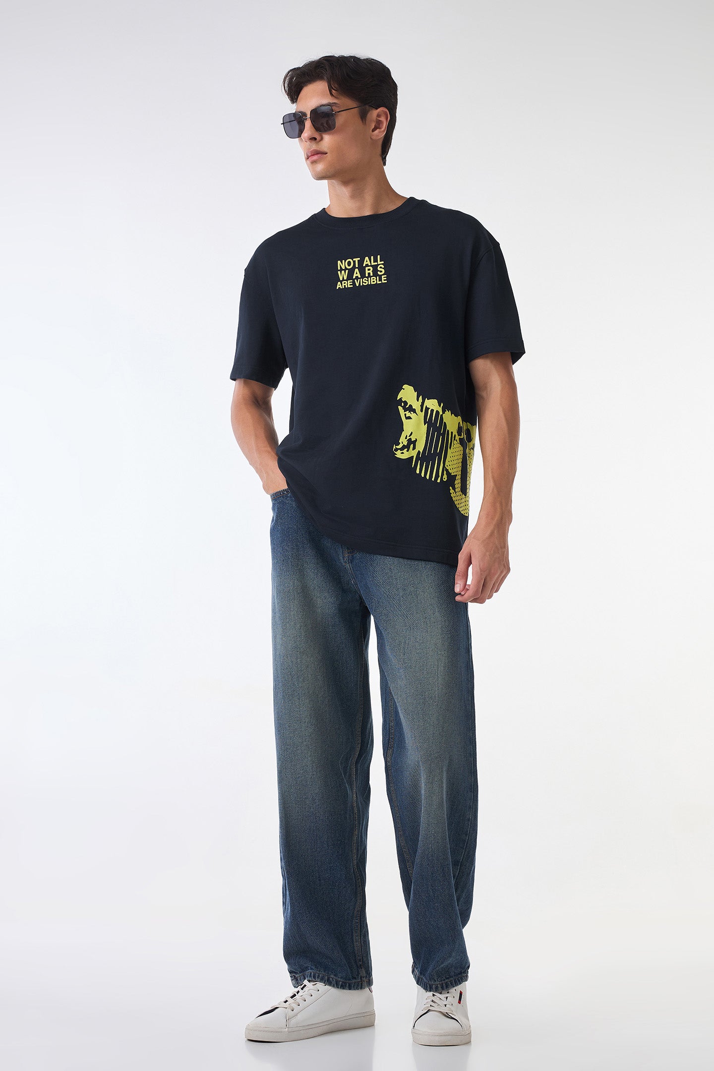 Full-body studio shot of the same black and yellow statement t-shirt styled with relaxed blue jeans and white sneakers against a clean white background.