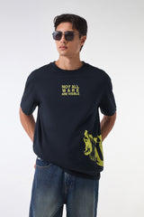 Front view of a man in a black statement t-shirt with yellow “NOT ALL WARS ARE VISIBLE” text and a bold yellow graphic near the hem, paired with blue jeans and hands in pockets.