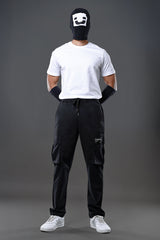 Man wearing black smiley mask, white t-shirt, black drawstring UNDERRATED CLUB cargo pants, and white sneakers against grey background.
