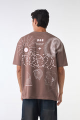 Back studio view of a man wearing a brown “888 Underrated Club” t-shirt covered with large white abstract graphics of a sun, stars, pixel patterns, and outlined heads, paired with dark jeans against a white background.