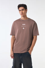 Front studio shot of a man wearing a brown “888 Underrated Club” graphic t-shirt with a small white star motif on the chest, paired with dark blue jeans against a clean white background.