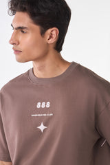 Close-up of a man wearing a brown t-shirt with minimalist white graphics on the chest, including “888,” “Underrated Club,” and a small star motif.