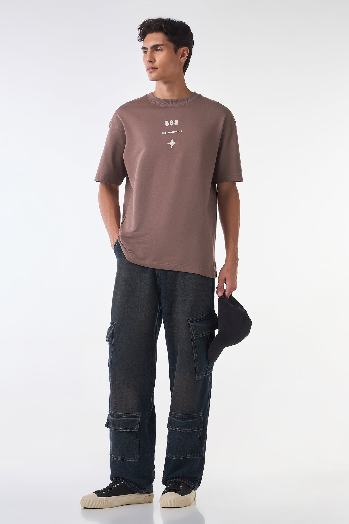 Full-body studio shot of the same brown graphic t-shirt styled with dark cargo jeans and sneakers, with the model holding a black cap in one hand against a white background.