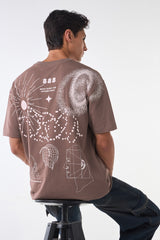 Seated back view of the brown t-shirt showing large white abstract illustrations of suns, stars, pixel patterns, and geometric faces, paired with dark jeans.