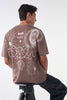 Seated back view of the brown t-shirt showing large white abstract illustrations of suns, stars, pixel patterns, and geometric faces, paired with dark jeans.