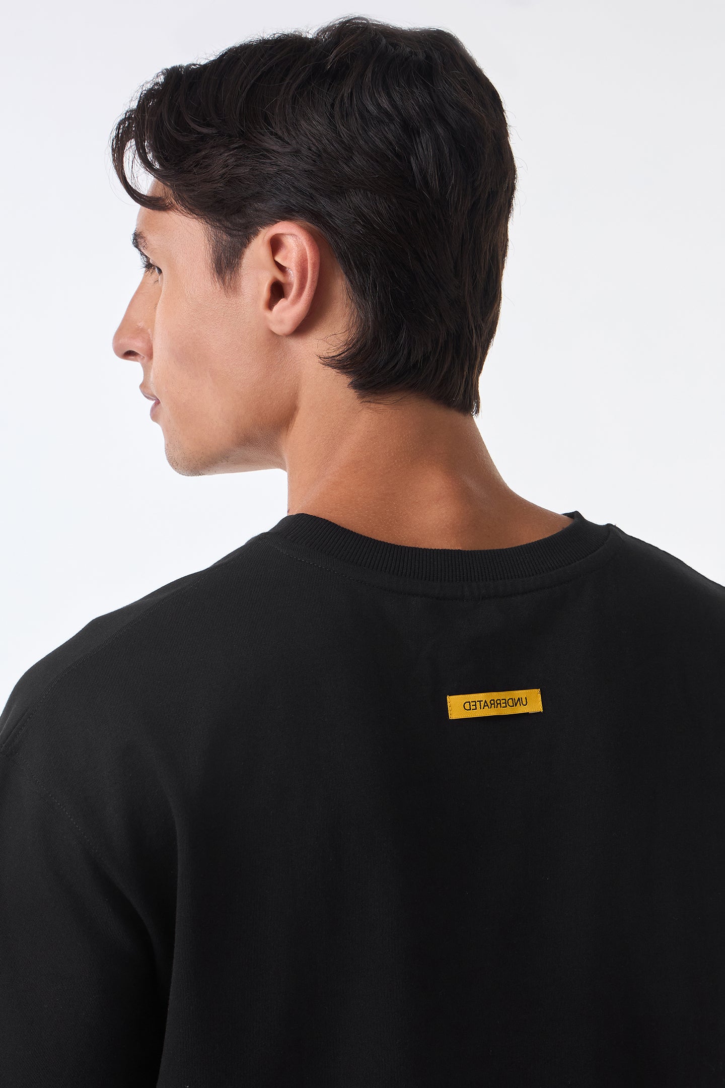 Man turning right shoulder view in black oversized t-shirt, arms relaxed, studio lighting.