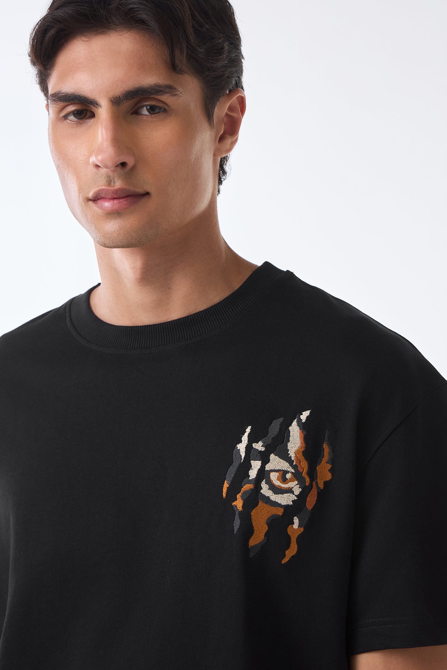 Close‑up of man's upper body wearing black crew neck t‑shirt with small chest graphic, neck cropped out, against plain background.
