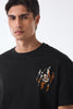 Close‑up of man's upper body wearing black crew neck t‑shirt with small chest graphic, neck cropped out, against plain background.
