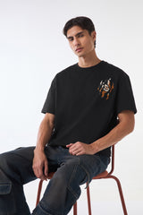 Man sitting on wooden stool wearing black oversized t-shirt, hands on knees, studio shot.