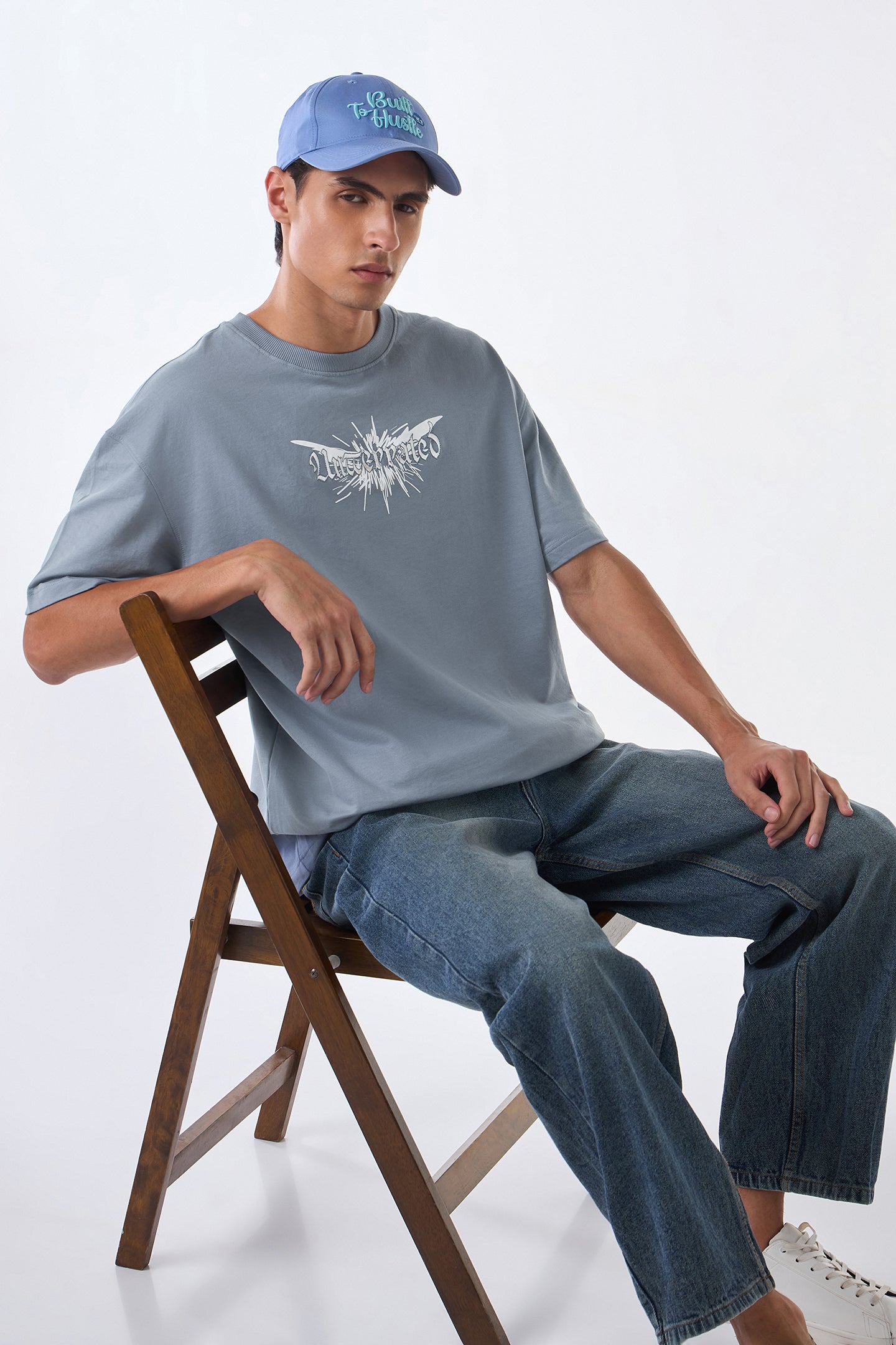 Man seated on wooden chair wearing light grey oversized t-shirt, cap backwards, legs crossed casually.
