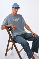Man seated on wooden chair wearing light grey oversized t-shirt, cap backwards, legs crossed casually.
