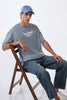Man seated on wooden chair wearing light grey oversized t-shirt, cap backwards, legs crossed casually.
