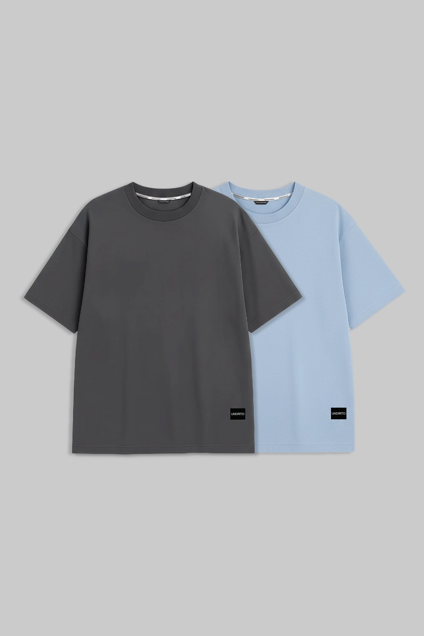 Two short-sleeve crewneck t-shirts displayed side-by-side against a solid gray background. The front t-shirt is dark charcoal gray, and the t-shirt behind it is light blue.