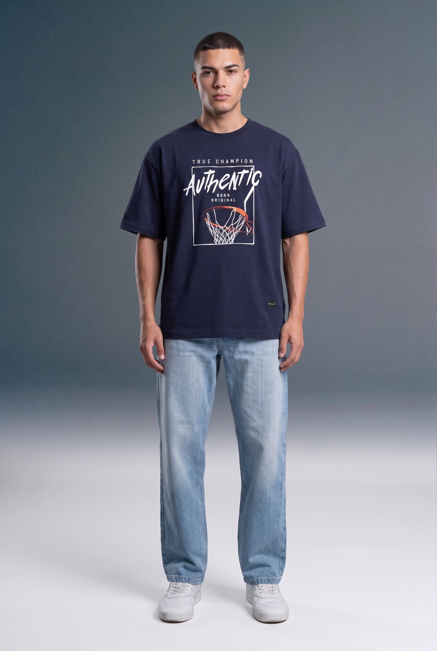 A man wearing a navy blue t-shirt with a white and red basketball graphic that reads 'AUTHENTIC CHAMPION', paired with light blue jeans and white sneakers, standing against a gray studio background.